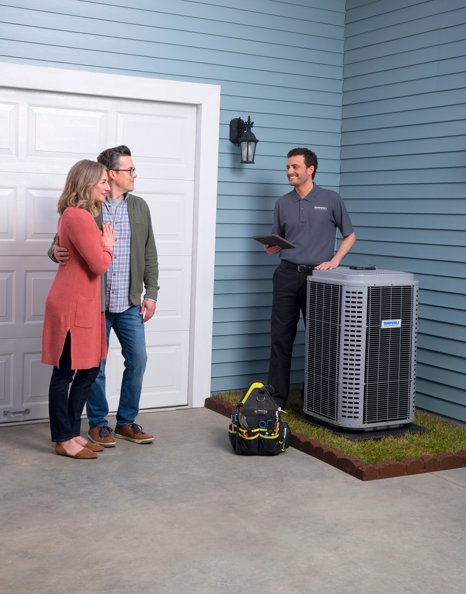 HVAC technician speaking with a smiling couple outside their home next to a central air conditioning unit, with tools on the ground nearby.